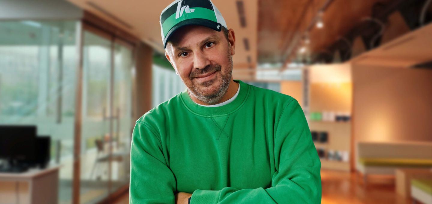 Person in einem modernen Büro steht mit verschränkten Armen. Die Person trägt ein grünes Sweatshirt und eine grün-weiße Cap. Im Hintergrund befinden sich helle, glasdurchzogene Arbeitsbereiche mit Schreibtischen und warmem Licht.