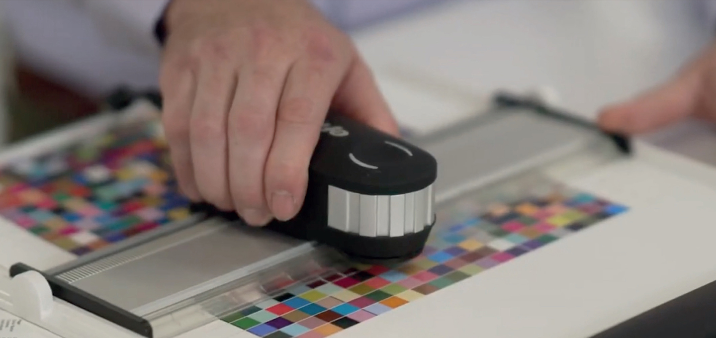 A hand uses a color calibration device on a sheet with vibrant color swatches. The focus is on technology and precision in printing or graphic design.