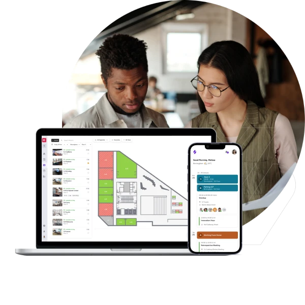 A person with light-colored hair, wearing a light-colored shirt, sits at a wooden desk, working on a laptop in a bright, modern office space. The office has large windows, plants, and a sign that says "RICOH Spaces." The person is looking at the laptop screen.