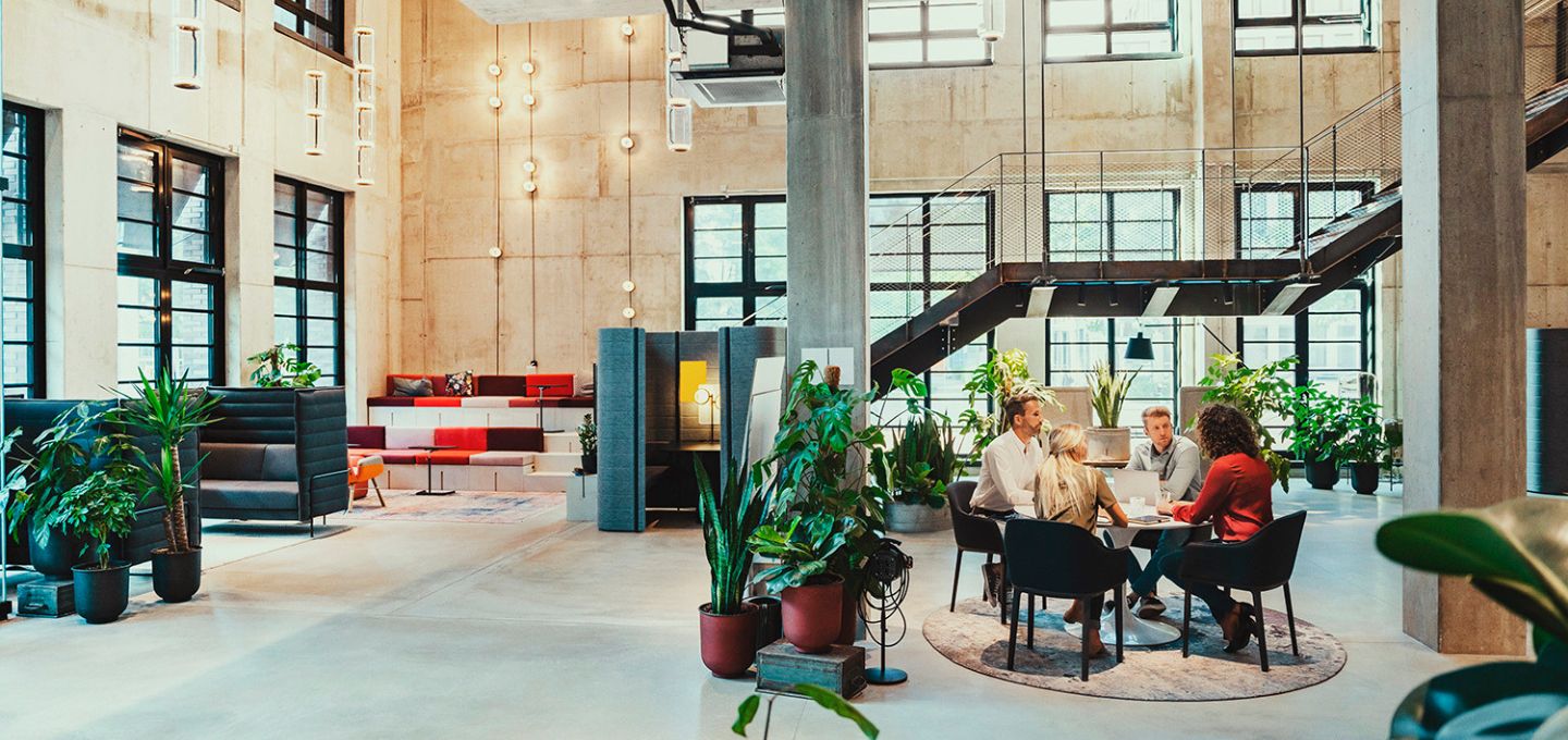 Modernes, offenes Büro mit Pflanzen, Sitzbereichen und einer Gruppe von Personen in einer Besprechung.